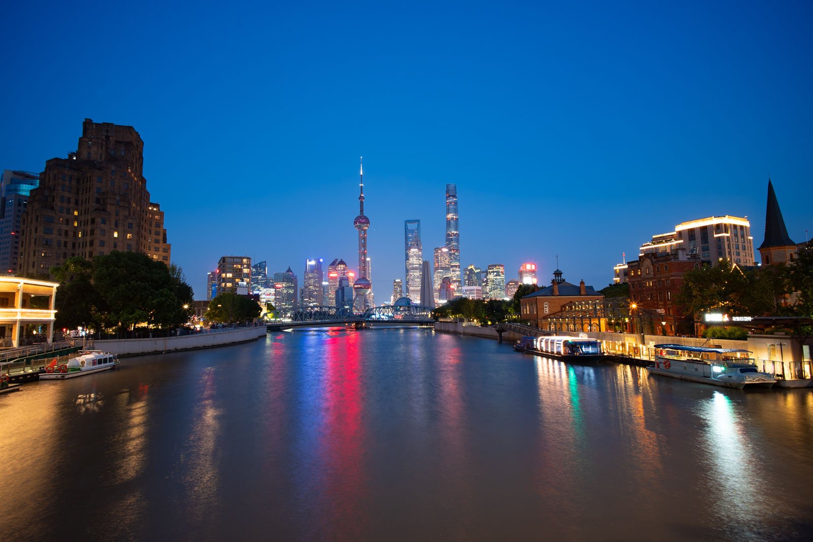 Night view of a vibrant city skyline with illuminated skyscrapers reflecting on a river. The sky is clear and deep blue, and the city lights create colorful reflections in the water. Boats are docked along the riverbanks.