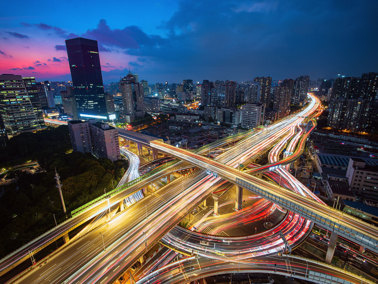 A vibrant cityscape at dusk showcasing a complex network of highways with bright streaks of car lights. Tall buildings surround the dynamic intersection, and the sky features a gradient of deep blue to pink as day transitions to night.