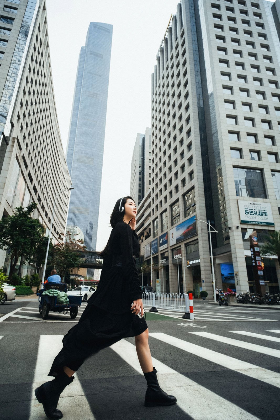 A person in a long black dress and boots walks confidently across a crosswalk in an urban setting, surrounded by tall skyscrapers and city traffic under a clear sky.