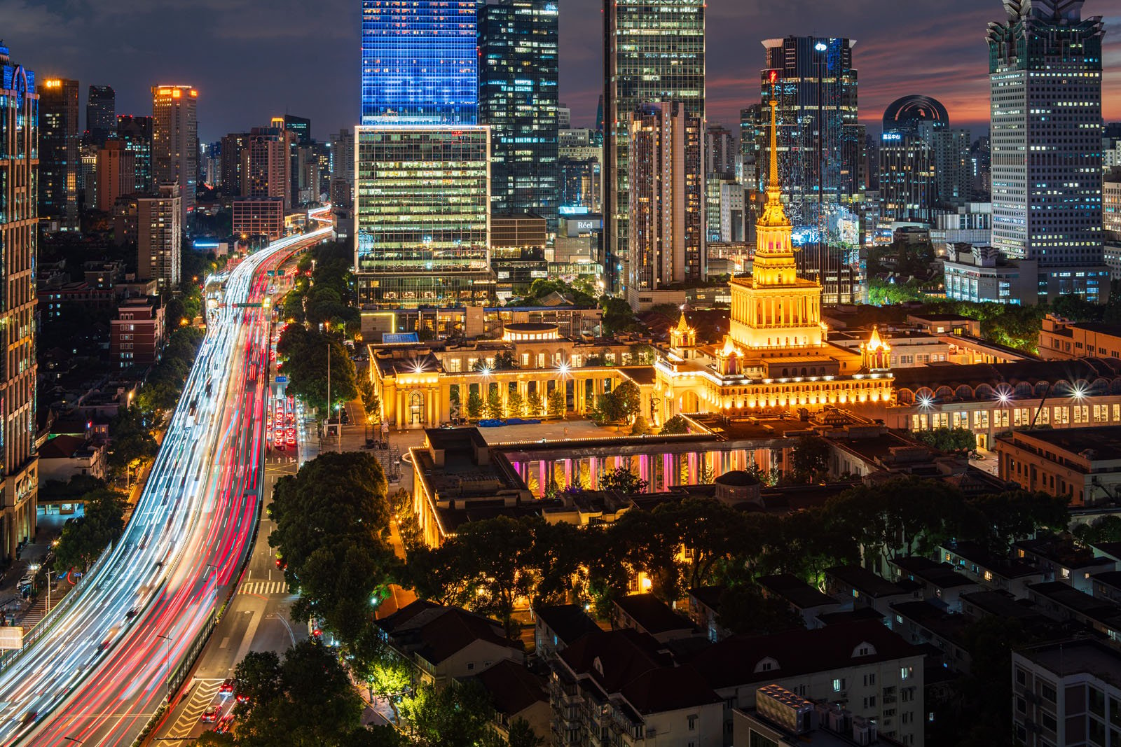 A nighttime cityscape featuring a brightly lit, ornate building with a spire in the foreground. Surrounding it are modern skyscrapers and a street with light trails from moving vehicles, indicating vibrant city life.