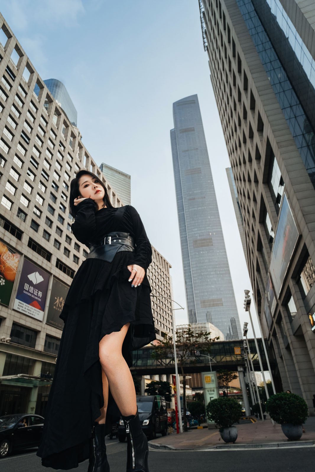 Woman in a stylish black outfit stands confidently on a city street, surrounded by tall skyscrapers under a clear sky.