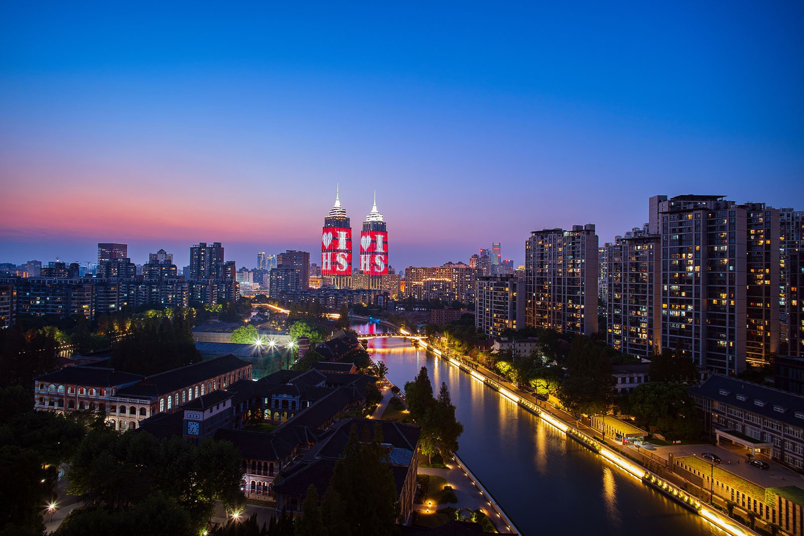 Cityscape at dusk with illuminated skyscrapers and a canal lined with buildings. The sky is a gradient of blue and pink, and the skyscrapers feature digital displays. Trees surround the waterway, adding to the urban landscape's tranquility.