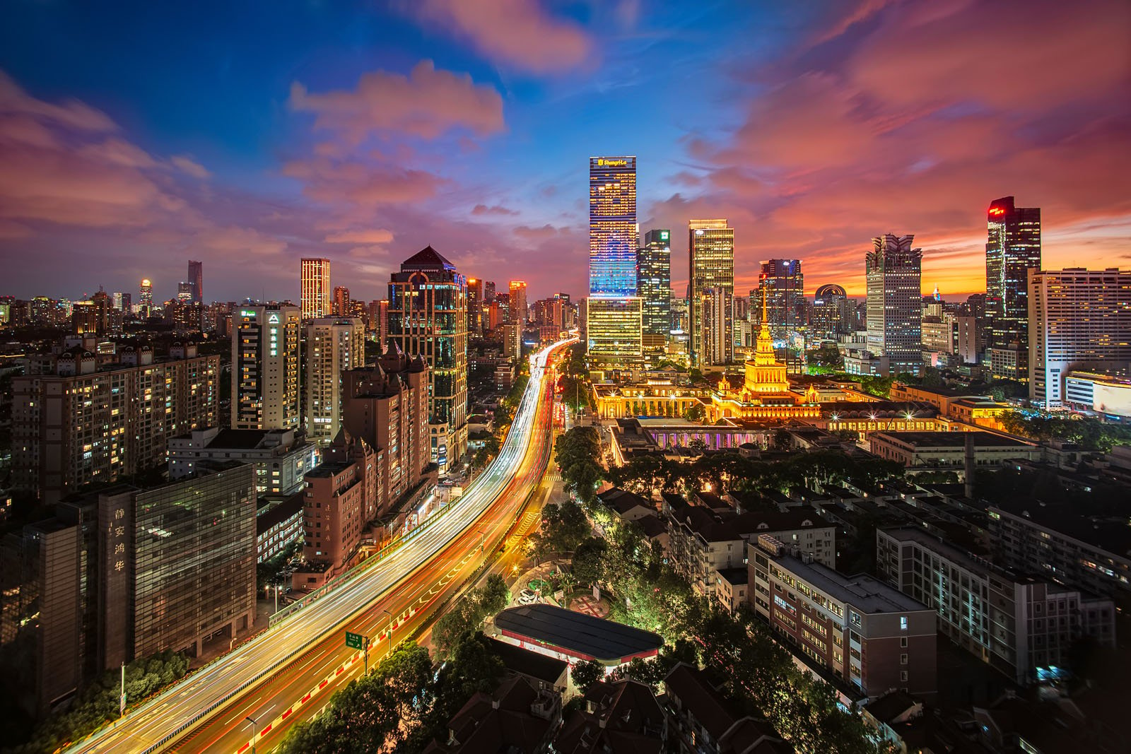 A vibrant cityscape at dusk, featuring tall, illuminated skyscrapers under a colorful sky. Bright streaks of light from traffic highlight a busy road leading towards the skyline, while the horizon glows with hues of orange and purple.