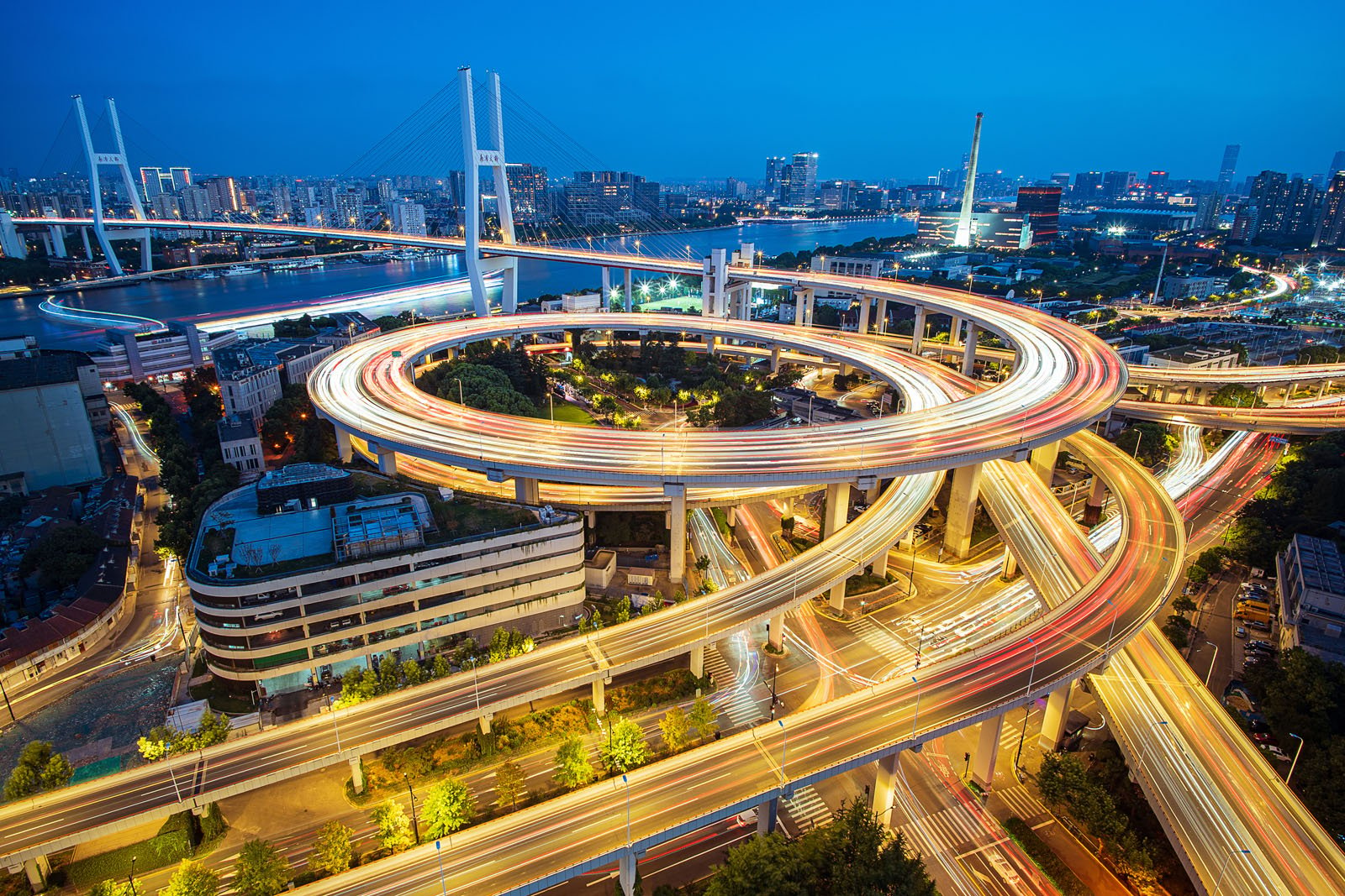 Aerial view of a vibrant city interchange at night, featuring multiple layers of winding roads and bridges. The long exposure captures the bright, colorful streaks of moving traffic lights, against a backdrop of a lit-up cityscape under a twilight sky.