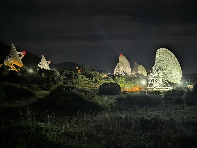 A collection of large radio telescopes is seen at night, illuminated by surrounding lights. The telescopes stand against a dark, starry sky with silhouetted hills in the background.