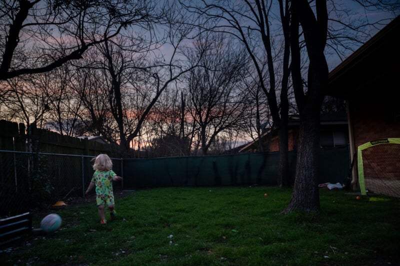 A small child in a colorful dress runs across a grassy yard at dusk, with a soccer ball nearby. Bare trees and a brick building surround the yard, while the sky is a mix of blues and pinks.