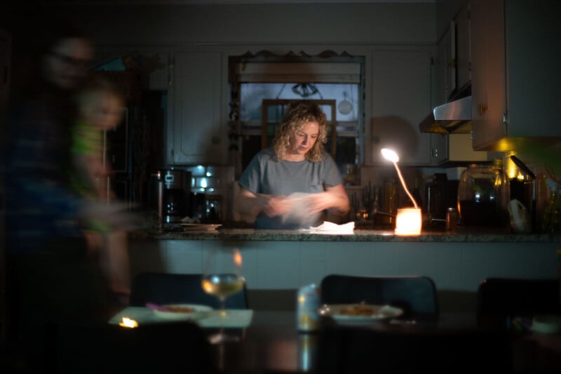 A woman stands at a kitchen counter in a dimly lit room, illuminated by a lamp. Blurry figures, including a person carrying a child, are seen in the foreground. Plates with food and drinks are on the table.