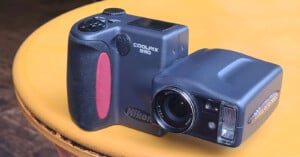 A Nikon Coolpix 990 digital camera with a unique swiveling lens is placed on a yellow circular surface. The camera is dark gray with red accents and has visible control buttons and branding.