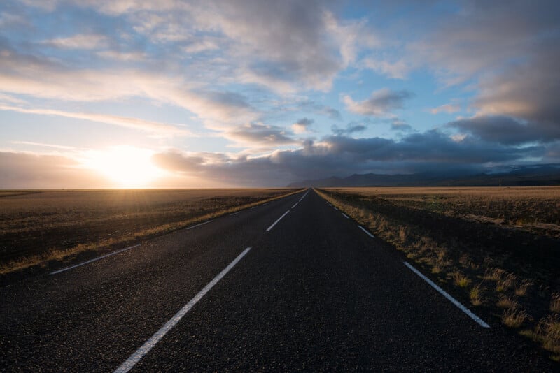 A long, desolate road extends toward the horizon beneath a partly cloudy sky. The sun is setting, casting a warm light across the landscape. Fields of grass flank both sides of the road, with distant mountains visible.