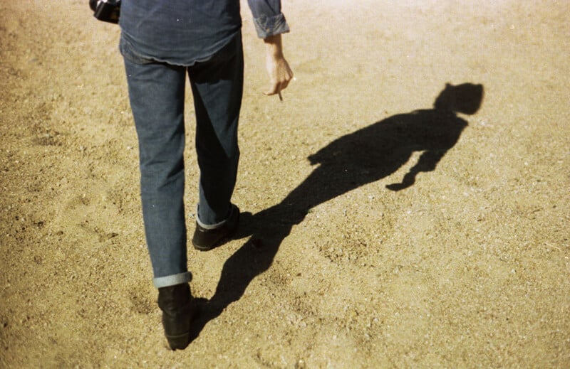 A person in jeans and boots walks on sandy ground, casting a long shadow. The shadow is distinct and elongated in the sunlight. The image captures motion and light contrast on the textured sand.