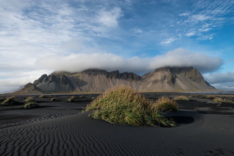 A scenic view of a black sand beach with green grass tufts in the foreground, leading to towering,⁢ cloud-topped mountains in the background. The sky is partly cloudy ​with patches of ​blue visible.