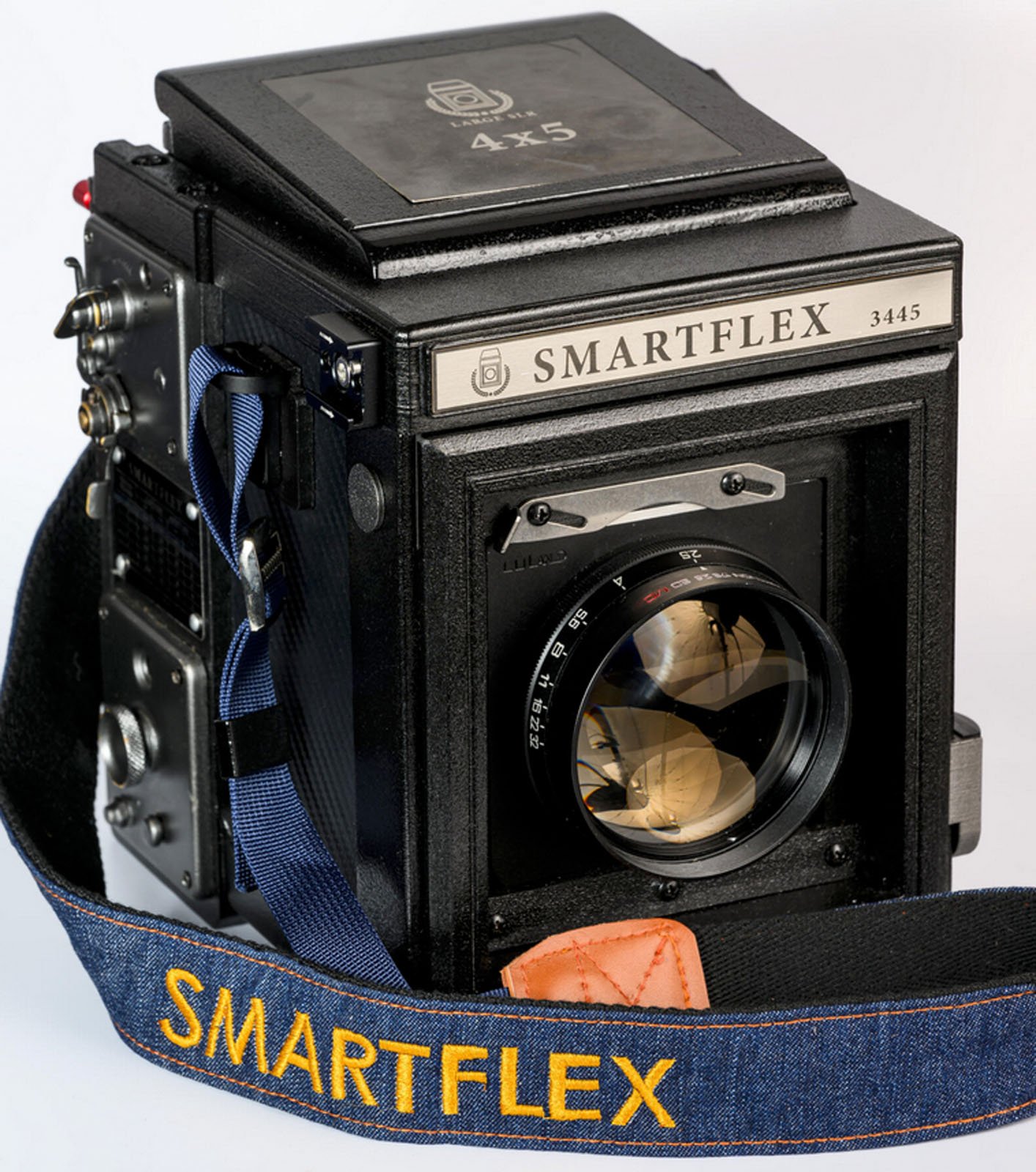 A vintage black box camera labeled "SMARTFLEX 3445" with a large lens on the front and a blue strap with "SMARTFLEX" written in yellow. The camera has a rectangular design with various controls on the side.