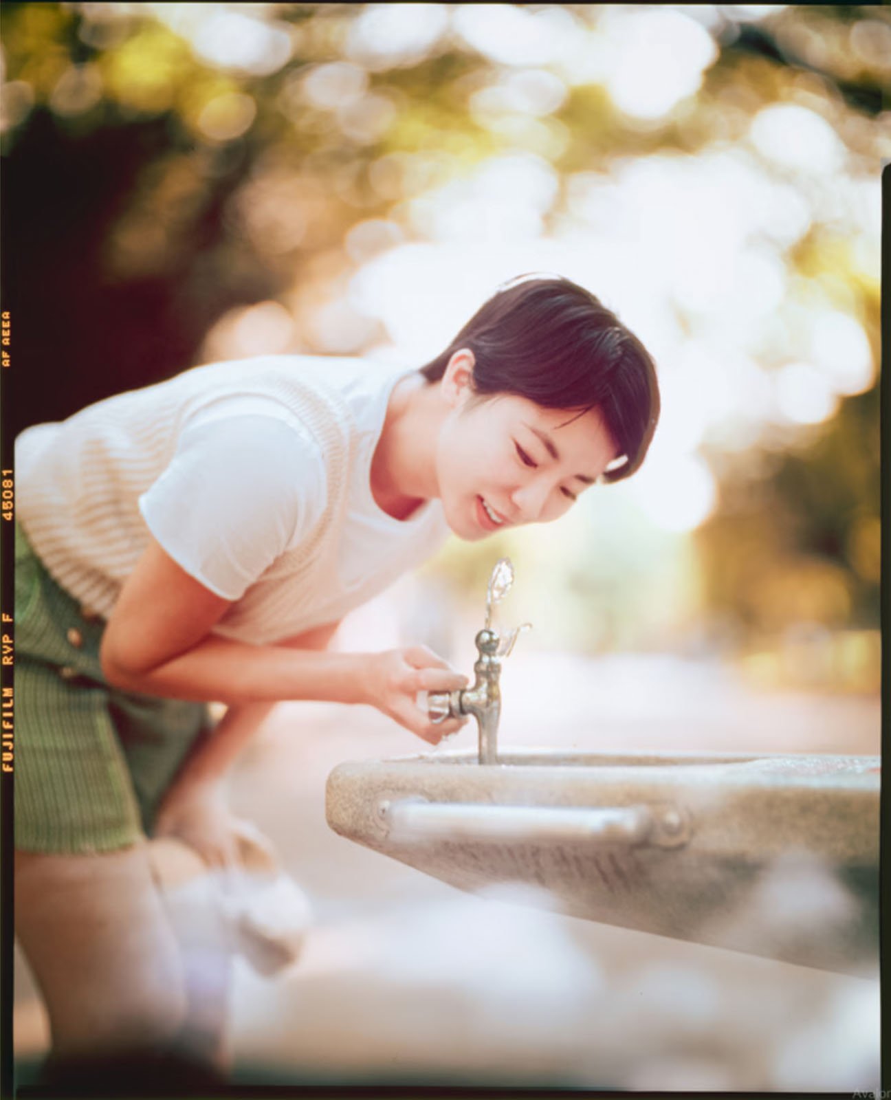 A person leans forward to drink from a water fountain outdoors. The background is softly blurred with greenery, suggesting a park setting.