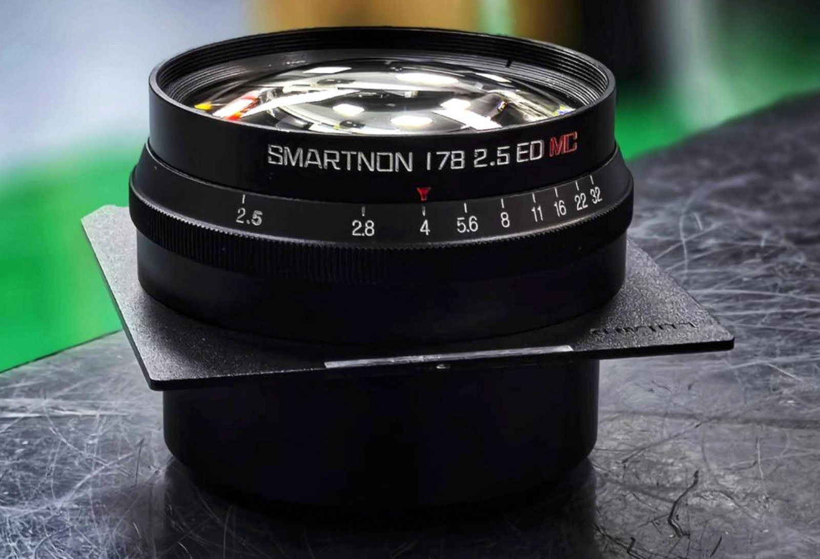 Close-up of a camera lens labeled "SMARTNON 178 2.5 ED MC" resting on a black surface. The background is softly blurred, highlighting the lens with a focus on its shiny glass and markings.