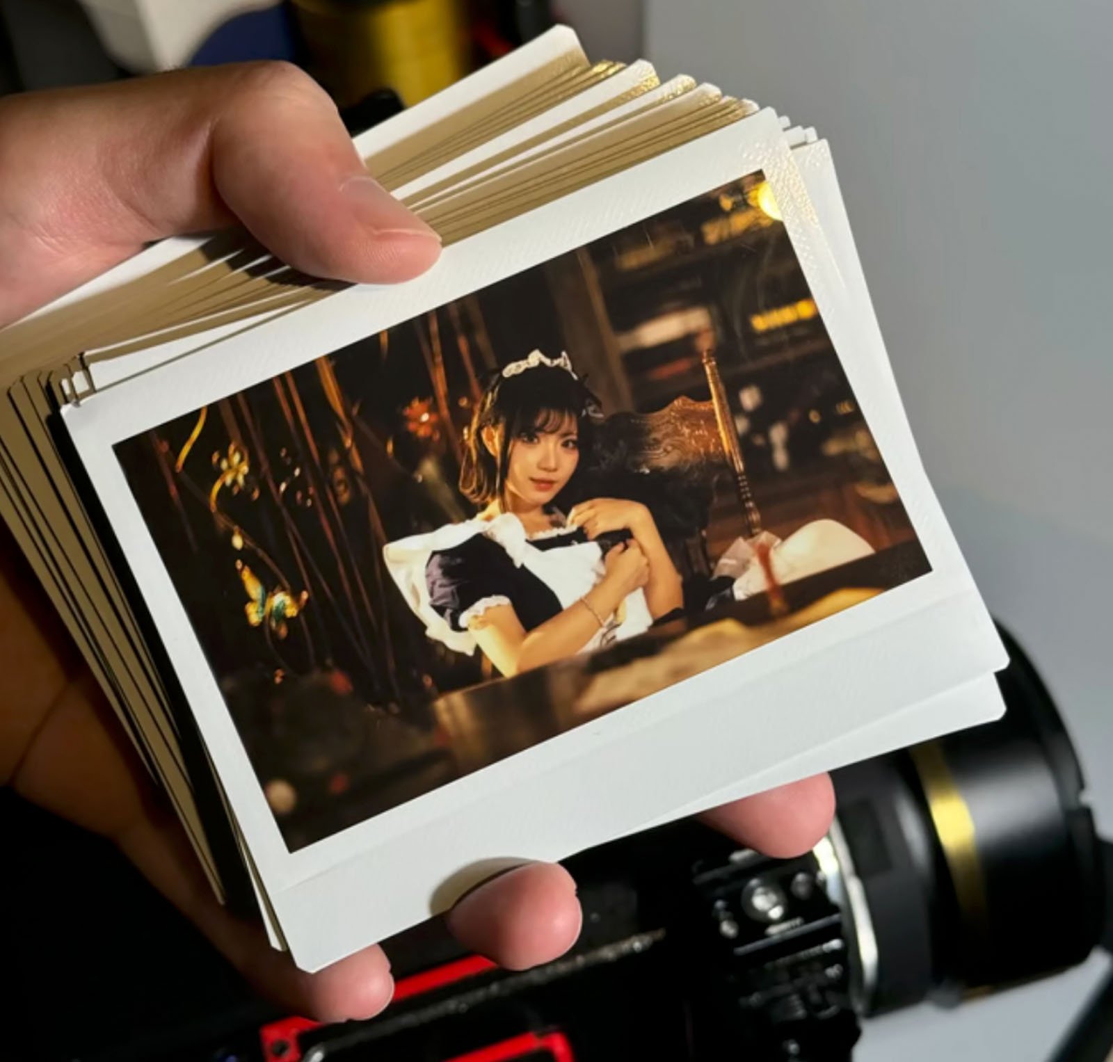 A hand holding a stack of instant photos, with the top photo showing a person in a maid costume, sitting in a dimly lit, ornate room filled with wooden furniture. A camera is visible in the background.