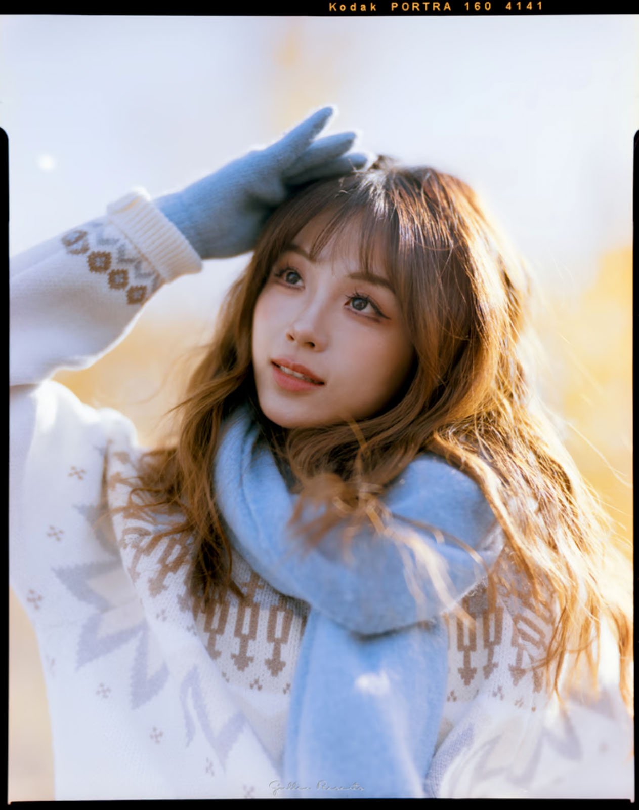 A woman in a white sweater and blue scarf and gloves gazes upwards in a sunny, outdoor setting. Her long hair flows around her shoulders, and the background is softly blurred in warm tones.