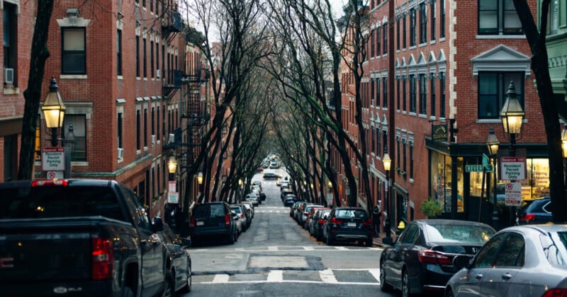 A street lined with red brick buildings and leafless trees, with cars parked on both sides. The road descends slightly, leading to a crosswalk and distant intersection. Street lamps and traffic signs are visible along the way.