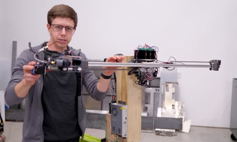 A person wearing glasses holds a long mechanical device with wires and components attached, standing in an indoor workshop. The background has various metal and wood materials.