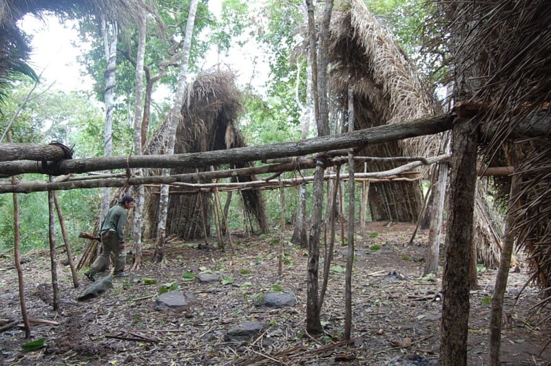 A person stands near a rustic wooden structure framed by logs and thatched roofs in a forest setting. The ground is covered in leaves and small rocks, and trees surround the area, suggesting a natural, rural environment.