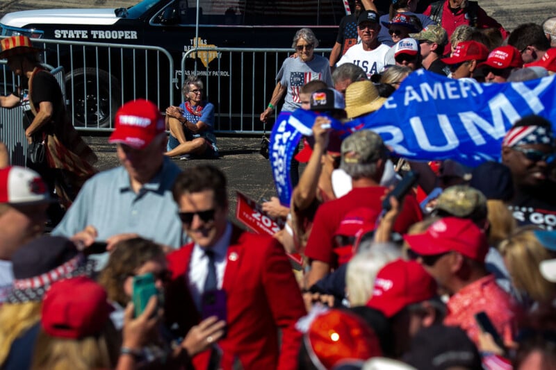 A crowd of people, many wearing red hats and holding a "Trump" banner, gather near a state trooper vehicle. A person sitting on the ground is visible in the background.