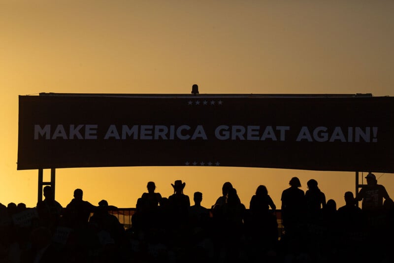 Silhouetted crowd in front of a large "MAKE AMERICA GREAT AGAIN!" banner at sunset. The sky is golden, creating a dramatic backdrop against the dark figures.