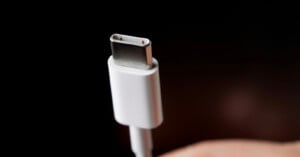 Close-up of a USB-C cable against a dark background. The focus is on the metallic connector at the end of the white cable, highlighting its distinct shape and design.