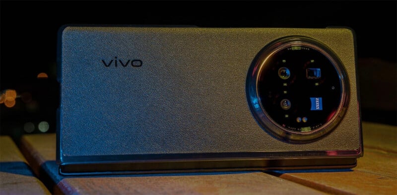 A Vivo smartphone with a textured back, placed on a wooden surface. The phone's camera module appears large and prominent, showing multiple lenses and a Zeiss logo. The lighting creates a dramatic effect against a dark background.