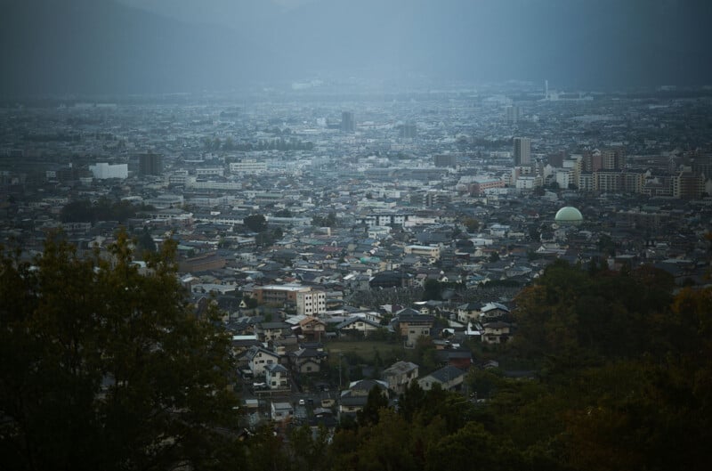 Aerial view of a sprawling cityscape on a cloudy day. Numerous buildings, houses, and streets are visible, set against a backdrop of distant mountains and a hazy sky. Trees frame the foreground.