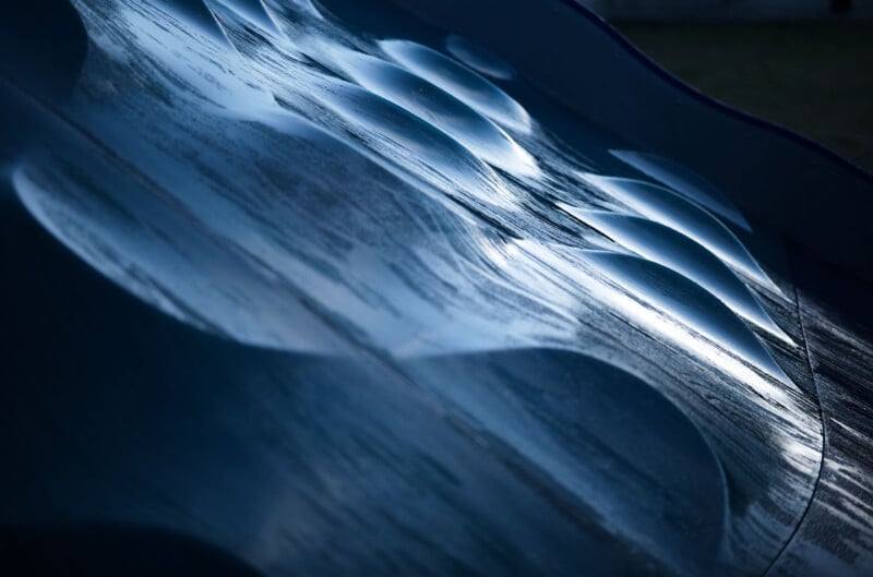 Close-up of an abstract metal surface with a textured, wavy pattern. The light reflects off the surface, creating a contrast between the illuminated and shadowed areas. The overall tone is cool and metallic.