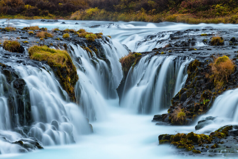 A serene waterfall cascades over dark, rocky ⁤ledges ⁤covered ⁣with patches of⁣ green moss and ‌grass. ⁤The water ⁢appears silky and ‍smooth, and surrounding ⁣foliage hints at an autumn setting with hints of yellow⁤ and ⁢brown.