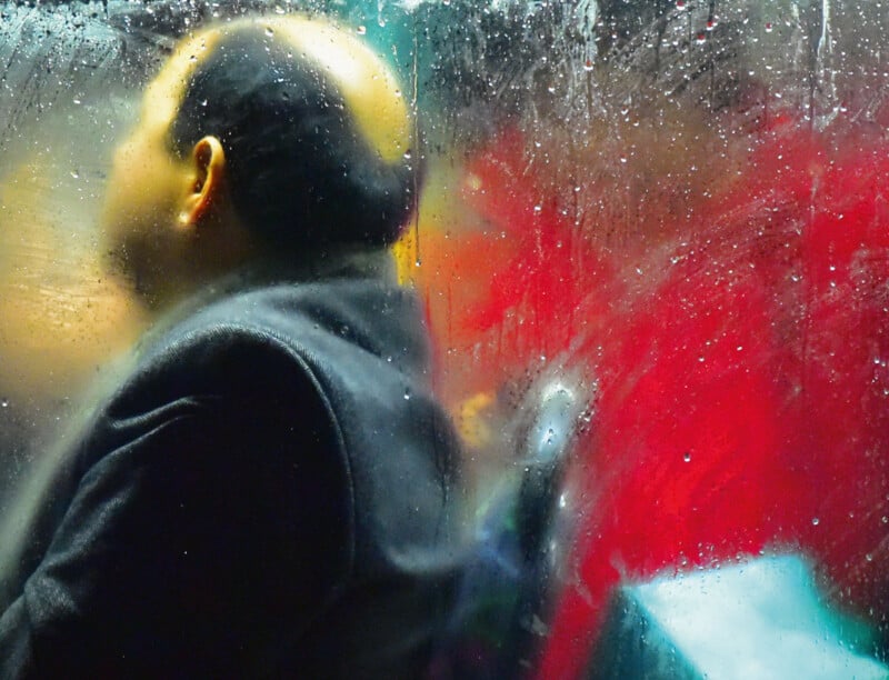 A person with a bald spot is seated and looking to the left, seen through a rain-covered window. The background is blurred with vibrant red and turquoise colors, creating a moody and abstract atmosphere.