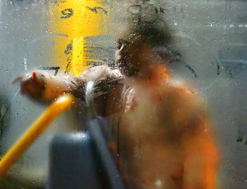 A blurred view of a person behind a wet, fogged-up window, with visible condensation and writing on the glass. The person appears to be pointing, set against a gray background with a bright yellow object inside.