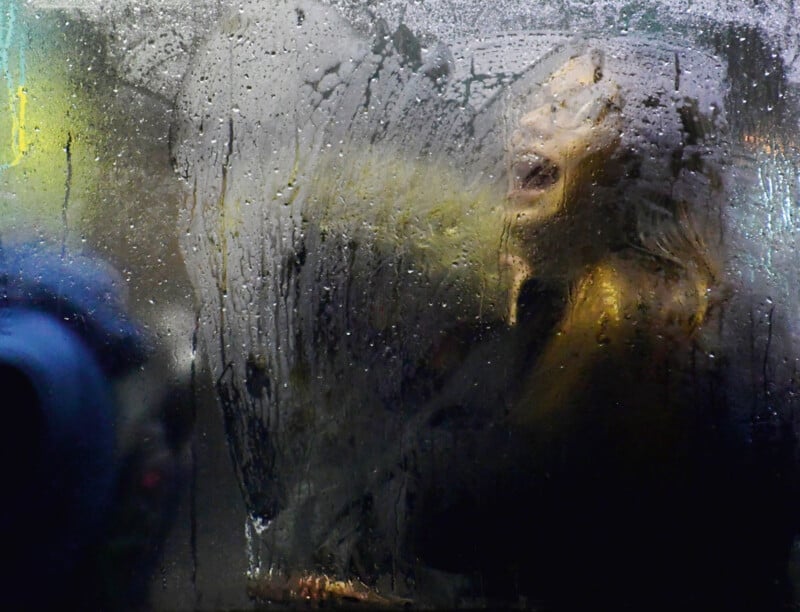 A person is visible through a foggy, wet window, creating a blurred effect. Their face appears expressive, with an open mouth, as if singing or shouting. The fog and condensation obscure the details, adding a mysterious atmosphere.