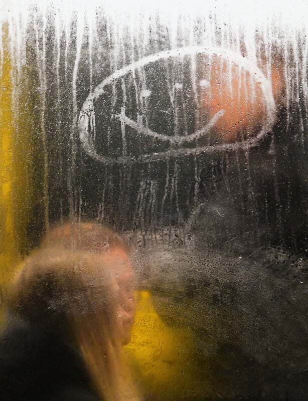 A foggy window with a smiley face drawn on it. Behind the window, a person is slightly visible, with their face blurred by condensation. The scene conveys a sense of warmth and mystery.