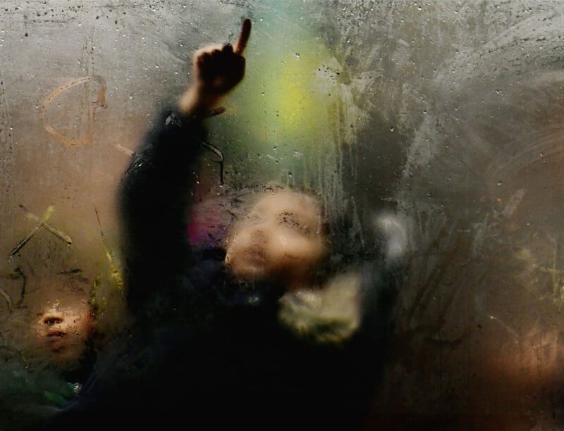 Two children are seen through a foggy, partially transparent surface. One child points upward, while the other looks on. The image is blurred, creating a mysterious and dreamy atmosphere.