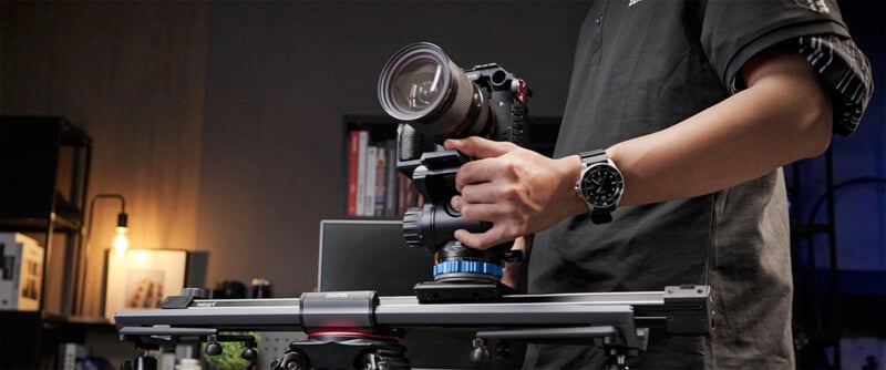 A person adjusts a professional camera on a rail slider in a dimly lit room. Various equipment and a soft light bulb are visible in the background. The person is wearing a black shirt and a watch.