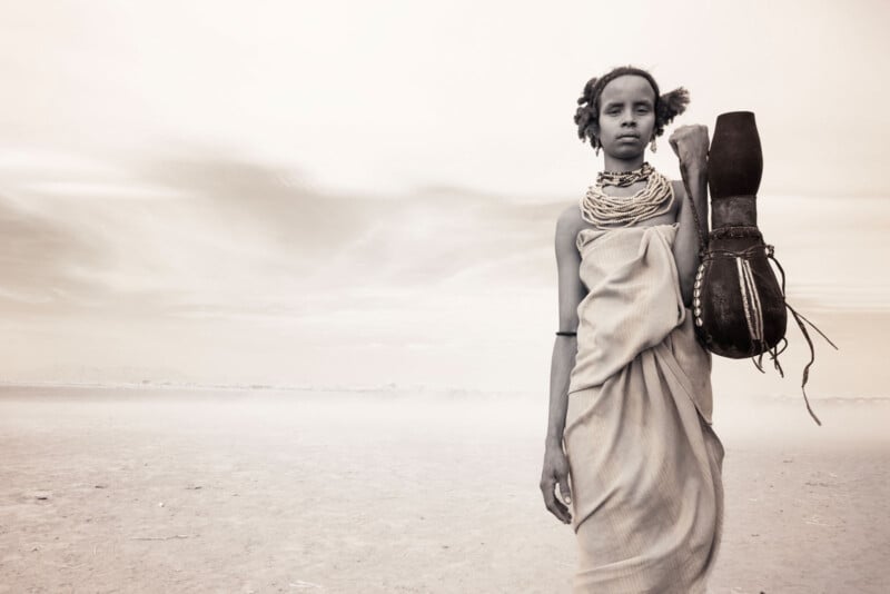 A person stands in a vast, arid landscape, holding a large vessel. They wear multiple necklaces and a wrapped garment. The background is hazy, and the scene appears monochromatic with a sepia tone.