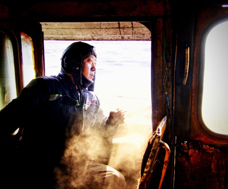 Person seated inside a rusty vehicle, wearing a helmet and jacket, looking out of an open door into bright light. Steam or smoke fills the space, creating a dramatic atmosphere.