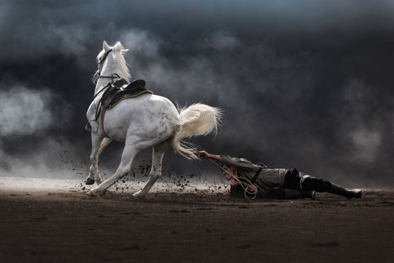 A person is being dragged on the ground while holding onto the tail of a galloping white horse. The scene is dramatic, with dust swirling around them against a dark, stormy background. The horse has a saddle but no rider.