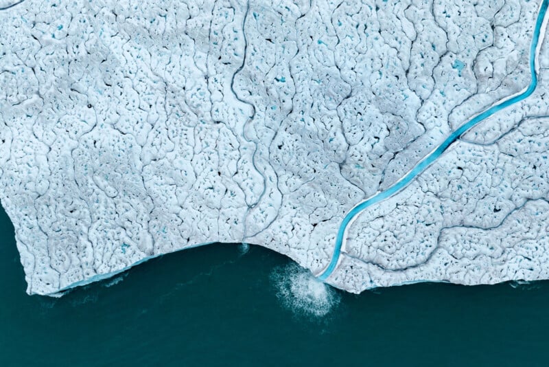 Aerial view of a melting glacier with crack patterns and a thin blue meltwater stream winding through it, leading to a body of dark blue water. Patterns resemble veins across the snowy surface.