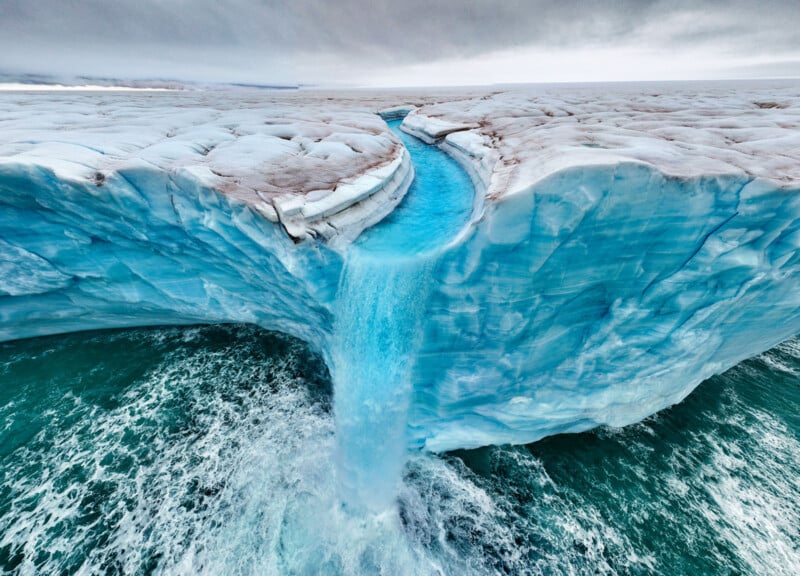 A stunning aerial view of a glacier showing a vibrant blue river cutting through and cascading off the edge into the ocean below, creating a dynamic waterfall amidst the icy landscape. The sky above is overcast, adding contrast to the scene.