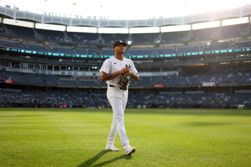 A baseball player in a striped uniform walks across the field under bright sunlight in a large, empty stadium. The sun sets in the background, casting long shadows on the grass.