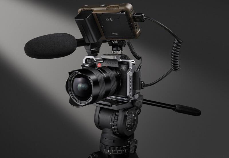 A professional camera setup featuring a mounted large microphone and an attached monitor. The camera is on a tripod, and the background is a neutral gray.