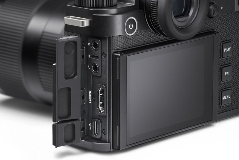 Close-up of a digital camera's side and back. The screen is off, and ports are visible on the side, including HDMI and USB. The camera body is black with textured grip, and several buttons such as Play and Menu are seen.