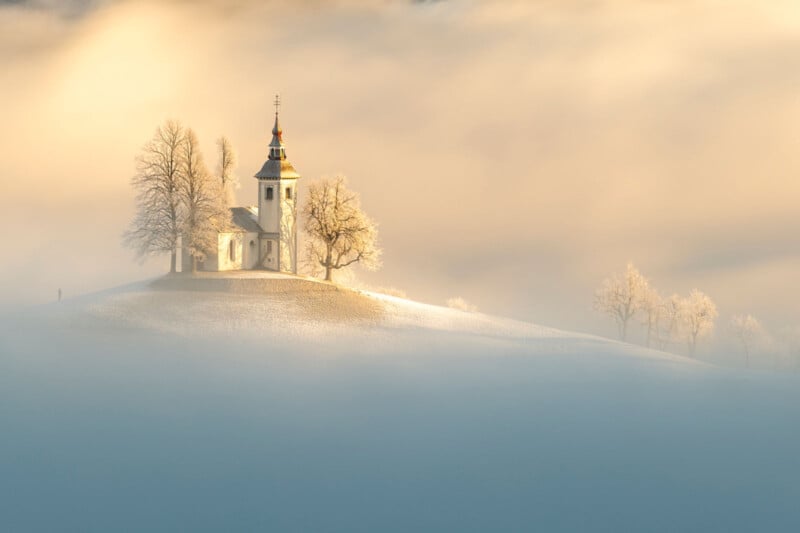 A small white church stands on a snowy hill surrounded by leafless trees. The scene is enveloped in soft fog, creating a serene and dreamlike atmosphere with a warm golden glow in the background.