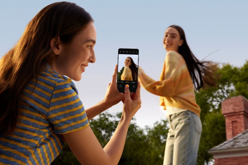 A woman in a striped shirt takes a photo with her phone of another woman smiling and posing playfully outdoors. The background shows trees and a roofline under a clear sky.