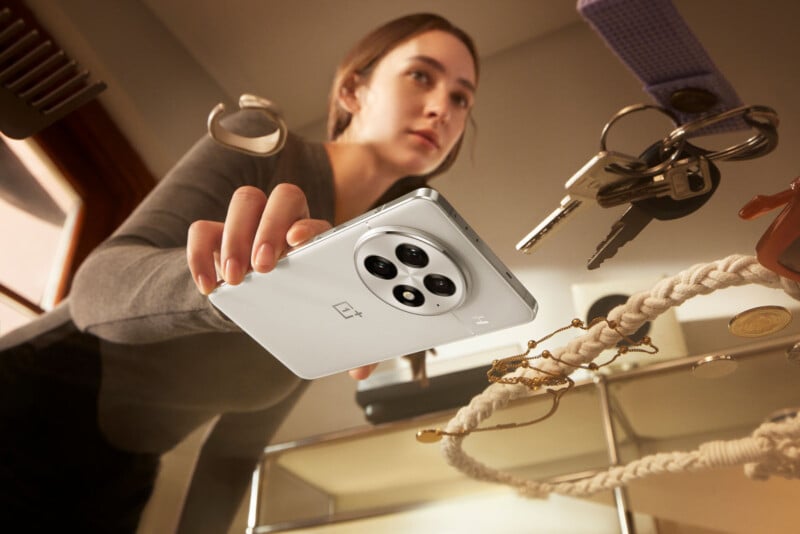 A person holds a white smartphone with a multi-lens camera, surrounded by floating objects including keys, a comb, and coins. The scene appears dynamic, as if the items are suspended in mid-air.