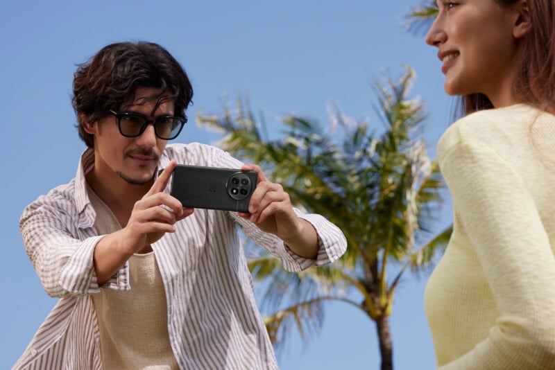 A person with spectacles capturing a photo with a smartphone of a joyous individual in yellow, set against a backdrop of a clear blue sky and palm treetops.