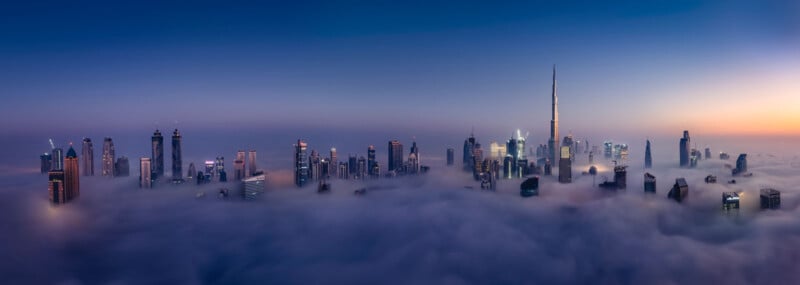 Panoramic view of a city skyline at dusk, with numerous skyscrapers emerging through thick, low-lying clouds. The tallest building stands prominently on the right, while the sky transitions from blue to orange as night falls.