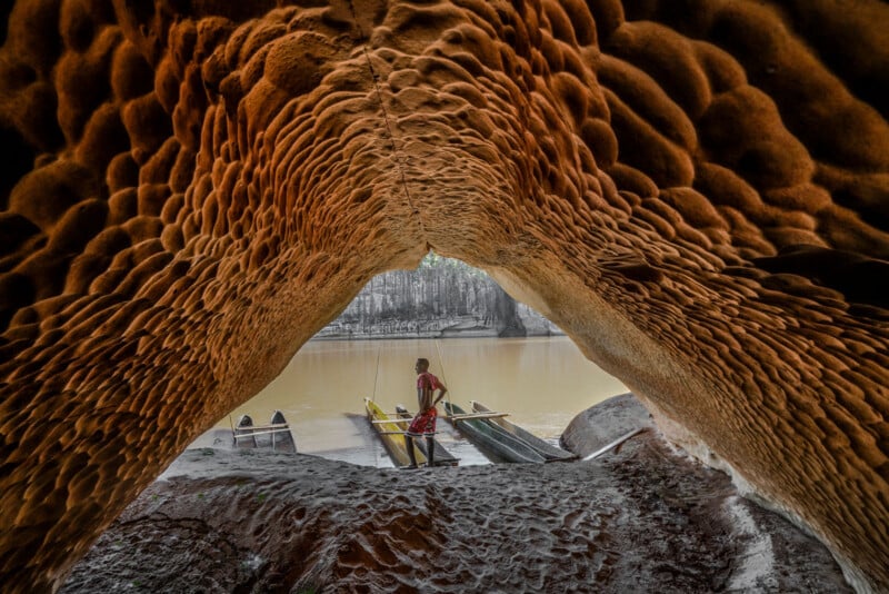 Inside a textured cave, a person stands on a wooden boat at the edge of a calm river. The view is framed by the cave's ceiling, revealing a serene outdoor scene with a forested background.